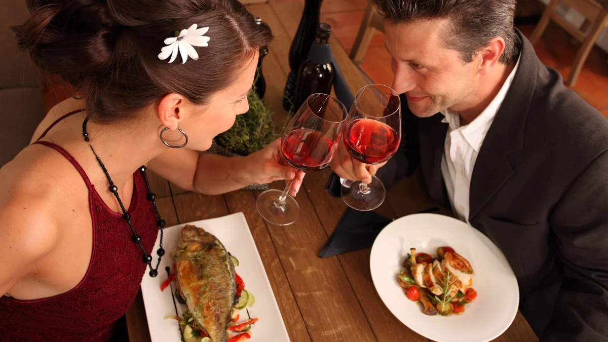 Mann und Frau sitzen im Restaurant gegenüber mit Essen vor sich und Wein in der Hand