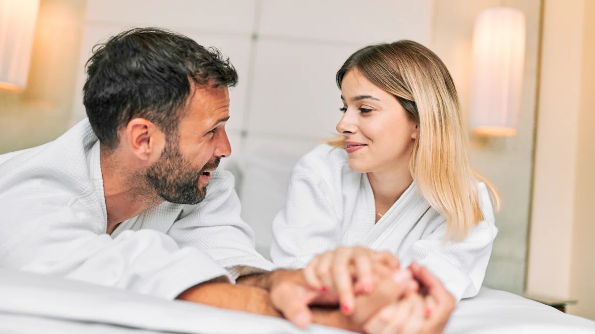 Man and woman laying in bed wearing a bath robe and holding hands